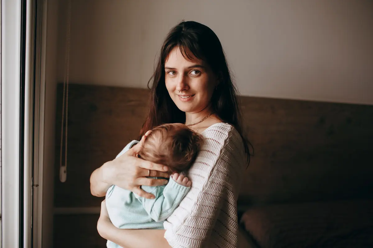 Matka drží v náručí spícího novorozence a stojí u okna v ložnici