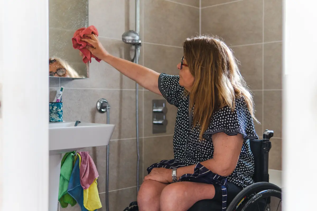 Žena na invalidním vozíku utírá zrcadlo v koupelně